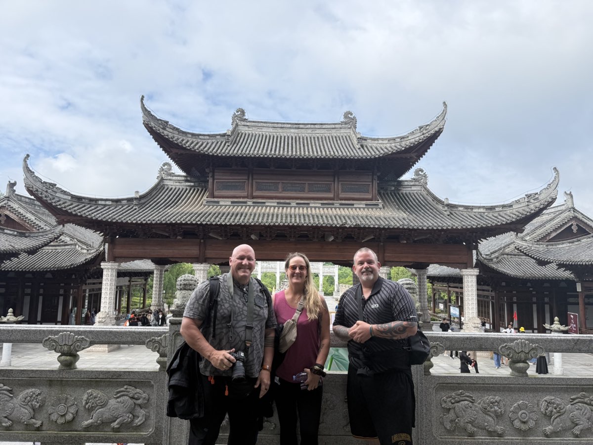 Three international guests smiling in front of a traditional temple building at Dazu